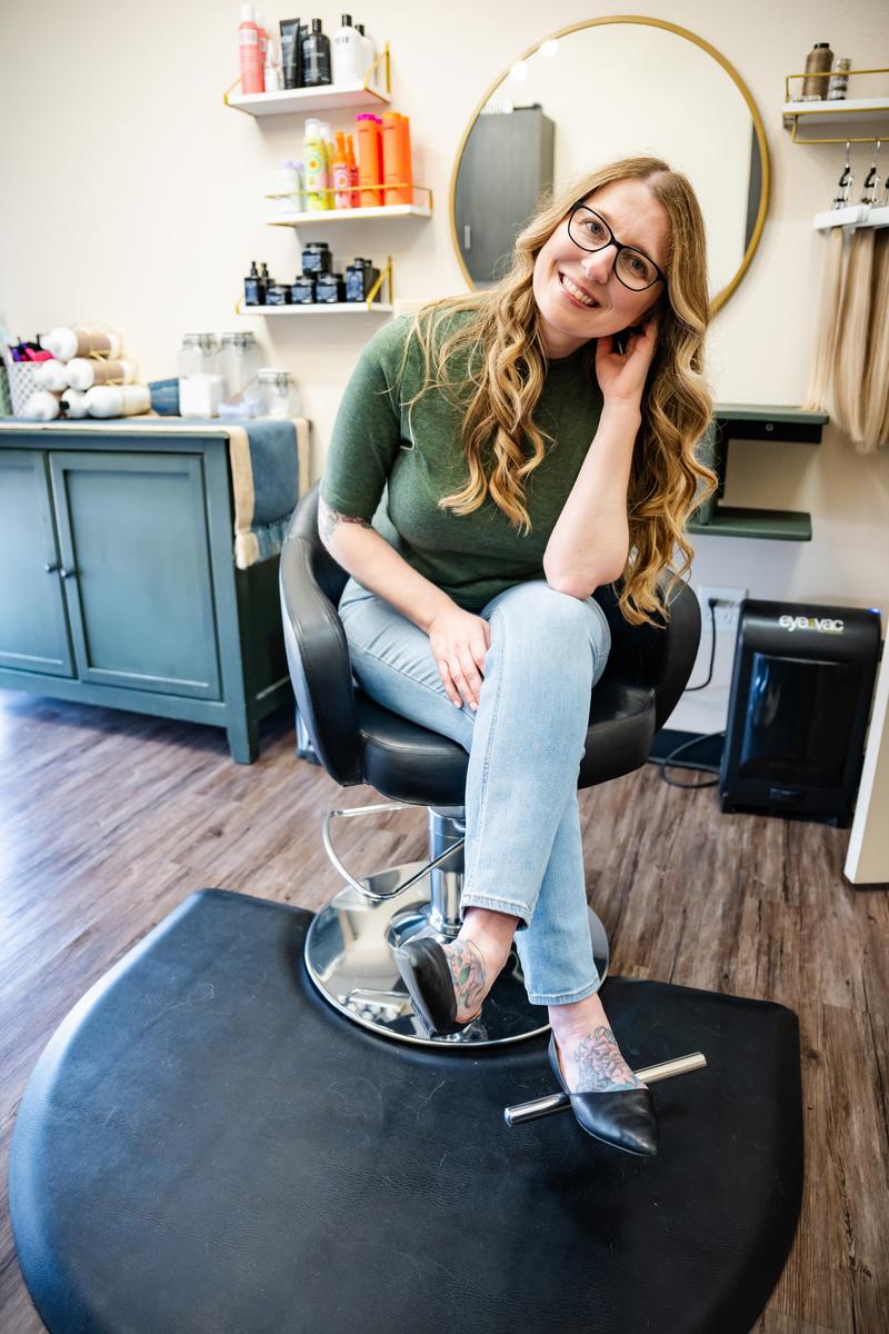 Blonde hairstylist smiling with her legs crossed wearing a green blouse sitting in her salon chair in Plano, TX.