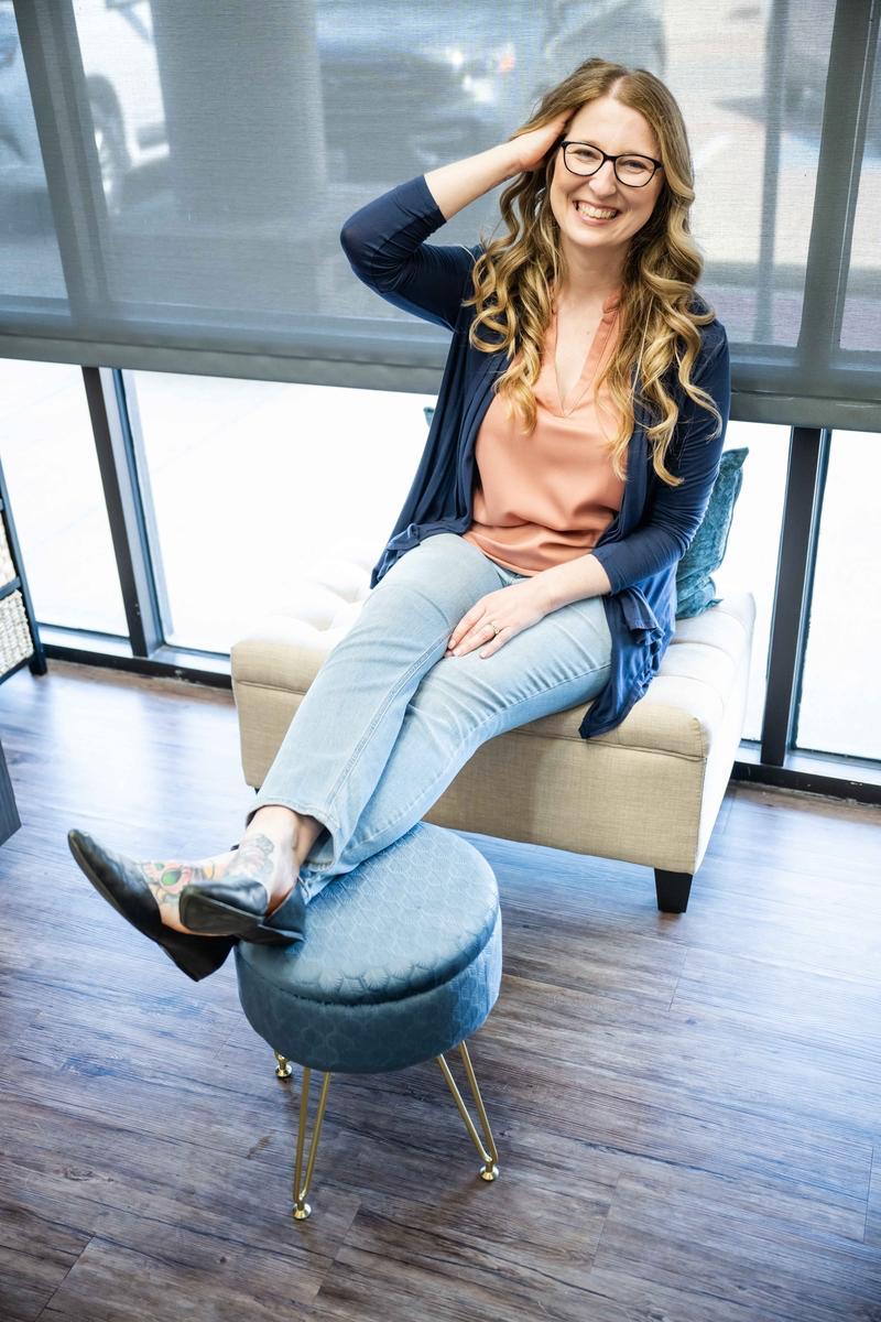 Hairstylist posing, smiling, with hands in her blonde hair wearing blue jeans and a blue cardigan in her salon in Plano, TX